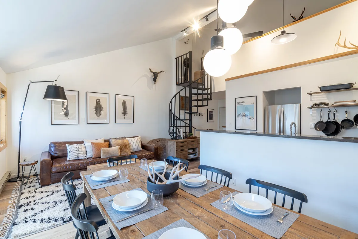 The hero image features a warmly lit open-concept living and dining area, with a stylish dining table set for a cozy meal, a plush sofa perfect for relaxing after a day outdoors, eye-catching wall art adding character, and a striking spiral staircase leading to a welcoming loft space. The ambiance is inviting, ideal for family gatherings or friendly celebrations.