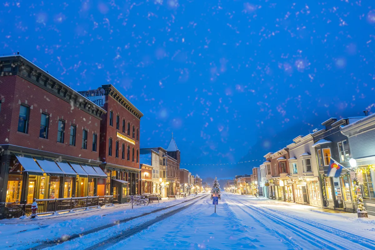 A snow-covered street in Telluride lined with historic buildings and festive decorative lights, featuring a large Christmas tree. The scene is tranquil, inviting families to stroll and enjoy local events.