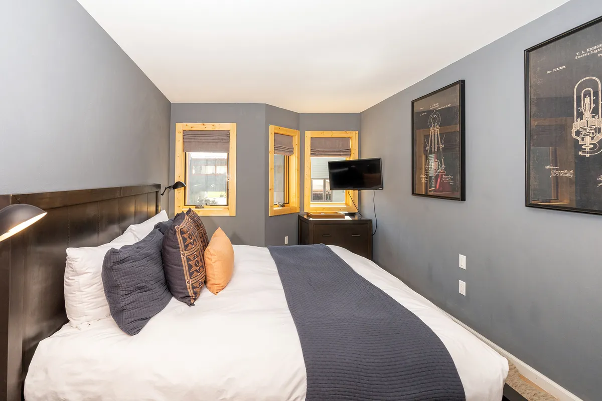 This image depicts a bedroom with a plush bed dressed in decorative pillows, framed artwork on the walls, wooden window frames, and a television on a stand. Soft natural light illuminates the space, creating an inviting, relaxing atmosphere ideal for restful mornings or winding down at night.