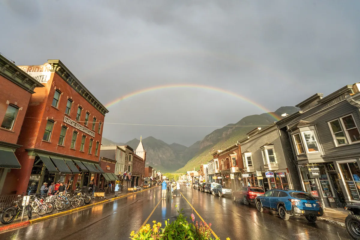 A picturesque view of Telluride’s historic main street with colorful old buildings, a rainbow after a rain, wet pavement reflecting the sky, and majestic mountains in the background. The scene conveys the vibrancy and charm of the town’s public spaces.