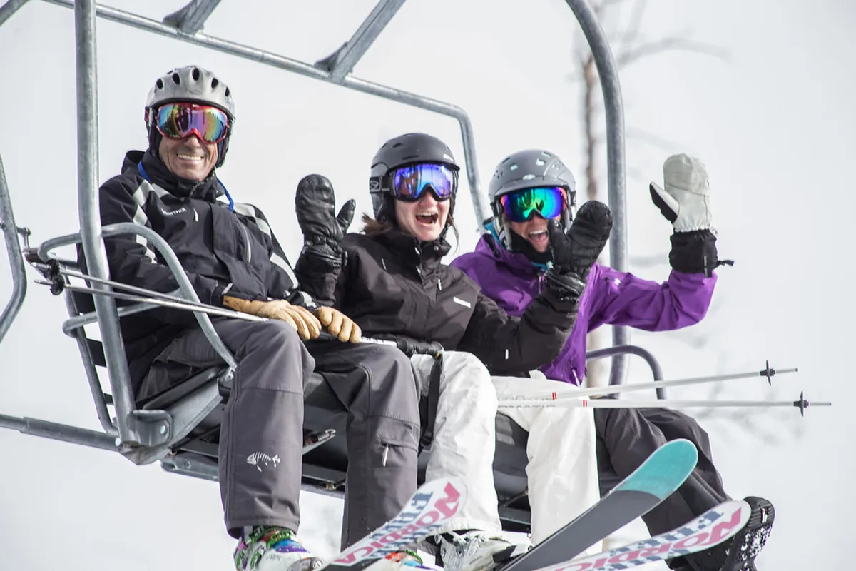 This image features three skiers riding a chairlift, surrounded by a snow-covered landscape with visible ski equipment and expansive mountain views. The playful mood captures the excitement and accessibility of ski adventures right outside the property.