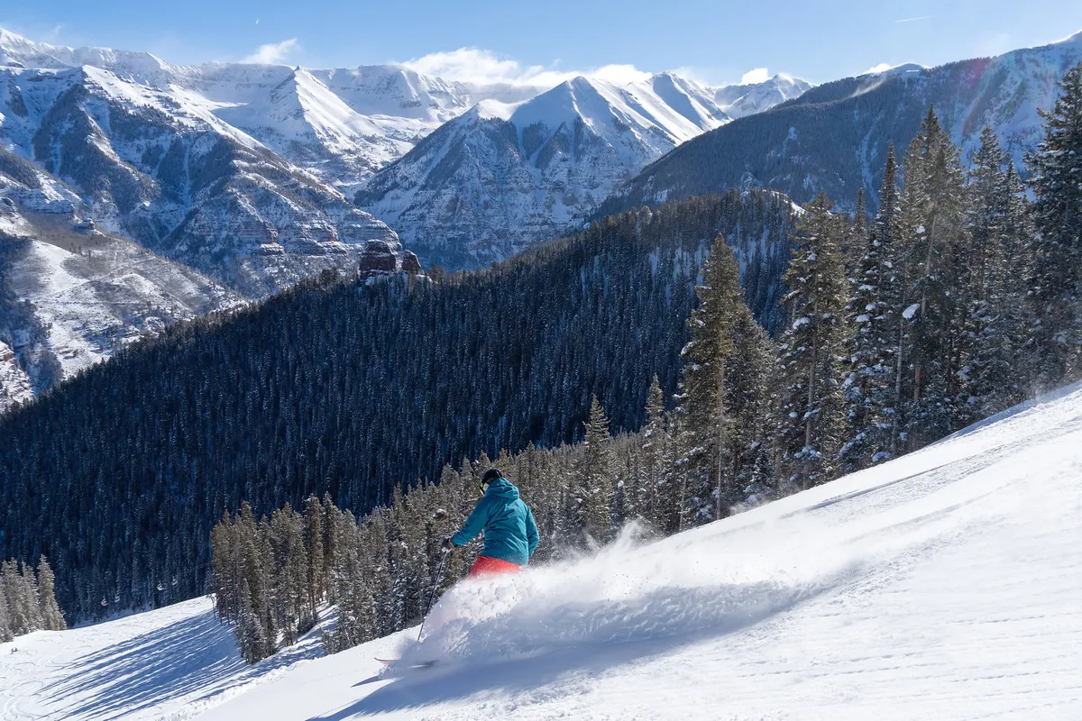 A skier glides across a snowy landscape framed by snow-covered mountains and evergreen trees. The action takes place under a clear sky, with an expansive view of the valley floor and the dramatic Telluride box canyon rising in the background.