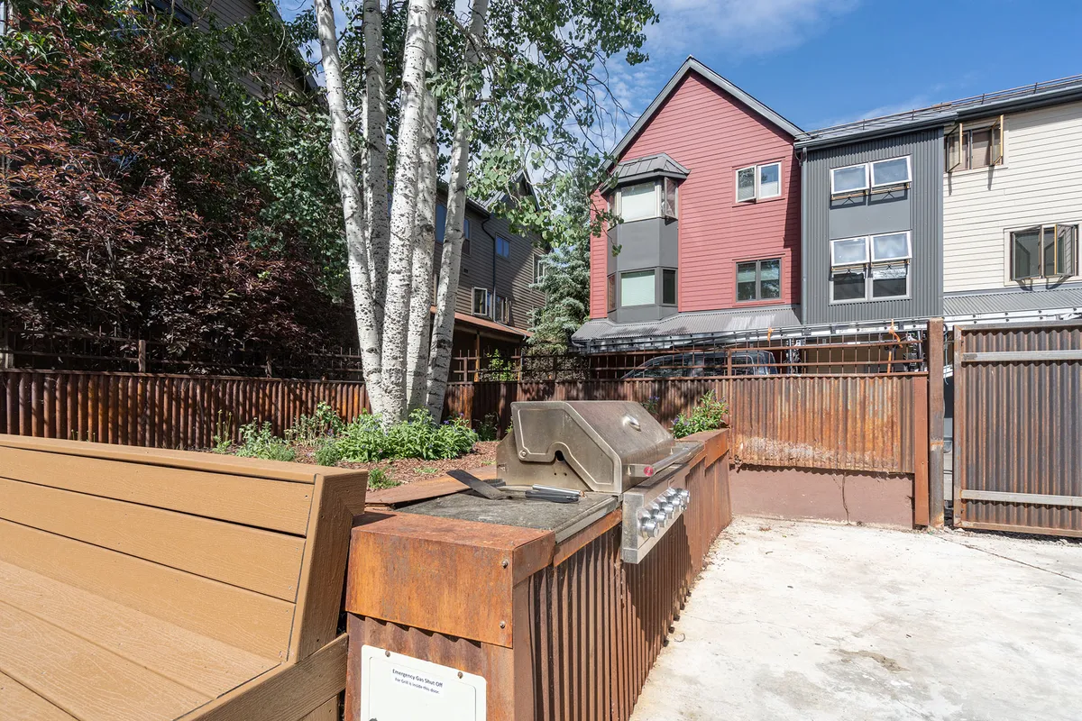 An inviting grill and seating area is nestled beneath leafy trees, adjacent to a building facade. The arrangement hints at leisurely summer evenings spent socializing, dining al fresco, or relaxing with friends and family.