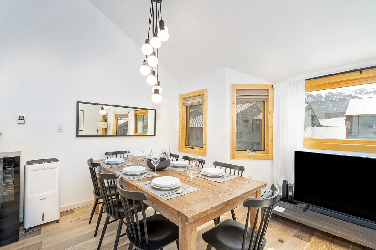A cozy and contemporary dining area with a stylish light fixture, a spacious table set for eight, and large windows that offer glimpses of the outdoors. The area is ideal for hosting special dinners and socializing after a day of exploration, further enhancing the communal atmosphere.