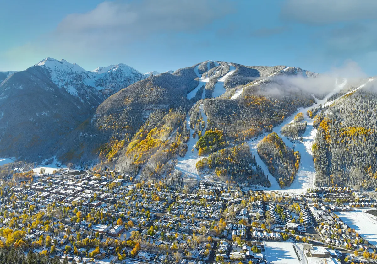 This exterior shot displays snow-capped mountains, visible ski slopes, the town nestled below, and autumn trees dusted with snow. The adventurous mood and scenic vista highlight both the natural beauty and lively winter atmosphere of Telluride.