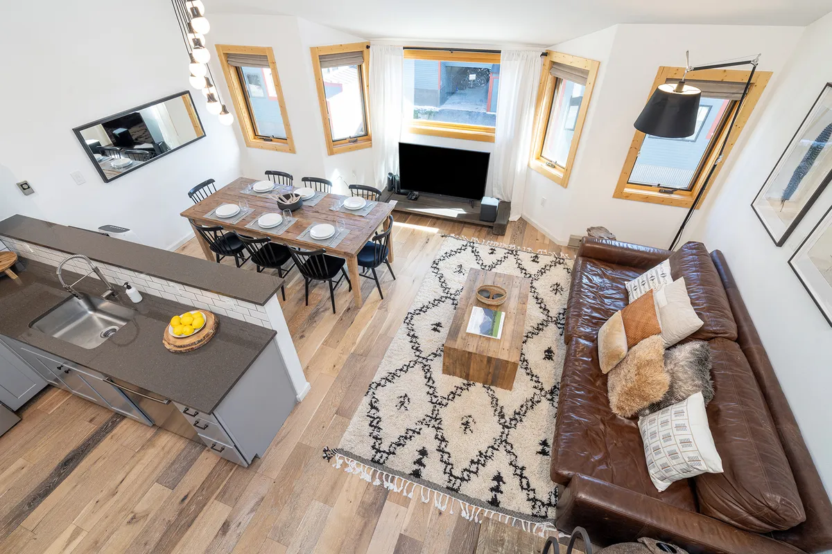 A bright, open-plan living and dining space featuring an oversized sofa, a large dining table with seating for eight, coffee table, TV, and expansive windows that flood the room with natural light. The view outside the windows hints at the scenic mountain surroundings. The layout encourages group gatherings, relaxation, and shared meals.