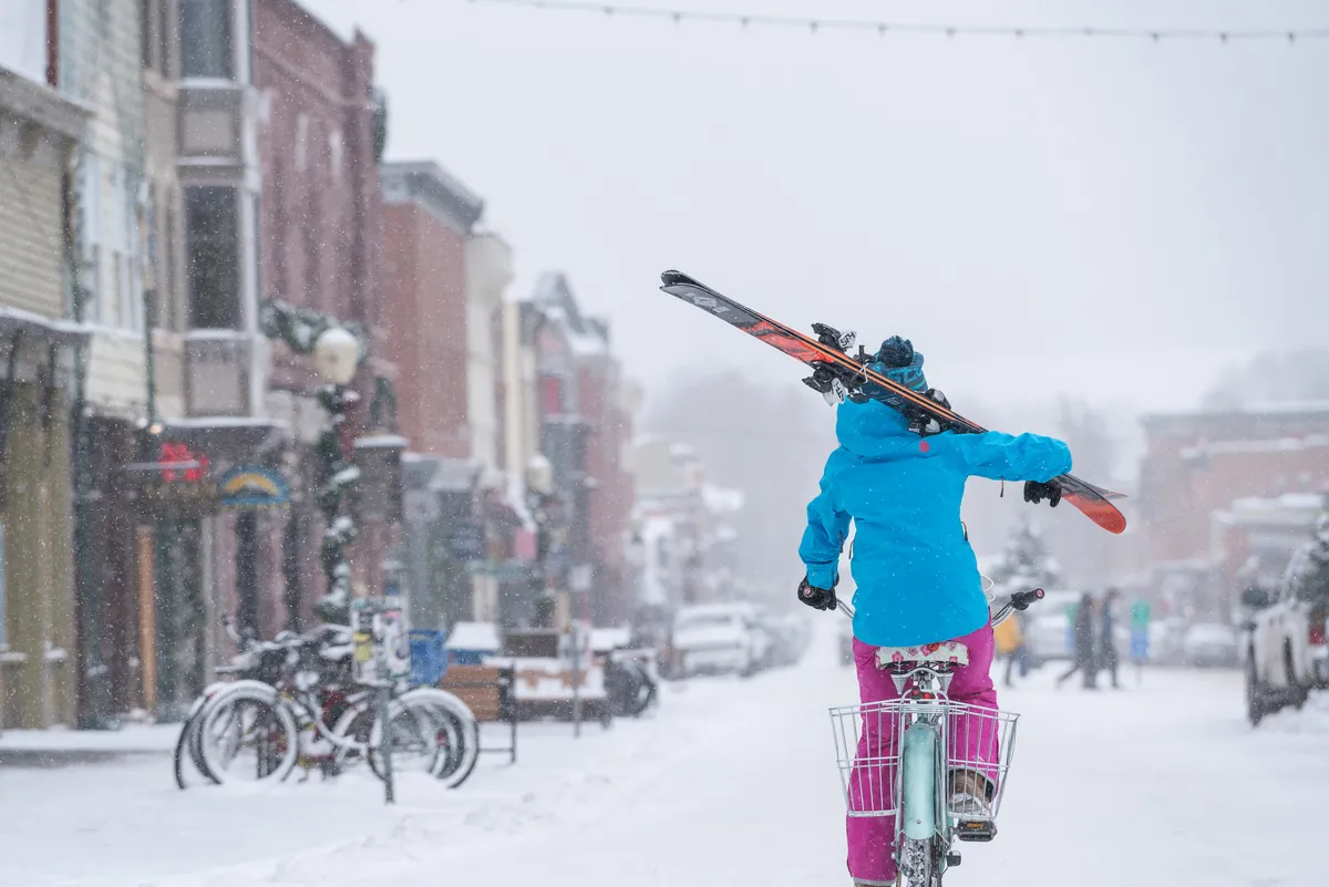 A snow-covered street in downtown Telluride with colorful storefronts lining either side. A person rides a bike while carrying skis over their shoulder, blending local adventure with easy access to the vibrant town. The atmosphere is lively, suggesting both activity and a welcoming community scene.
