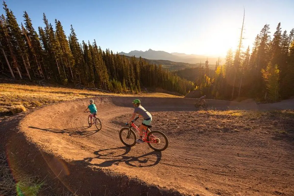 A group of mountain bikers navigates a curved dirt path at sunset, surrounded by pine trees and a rugged mountain range in the distance. The golden light of sunset highlights the vibrancy of the landscape, suggesting an atmosphere of outdoor adventure and summer activity.