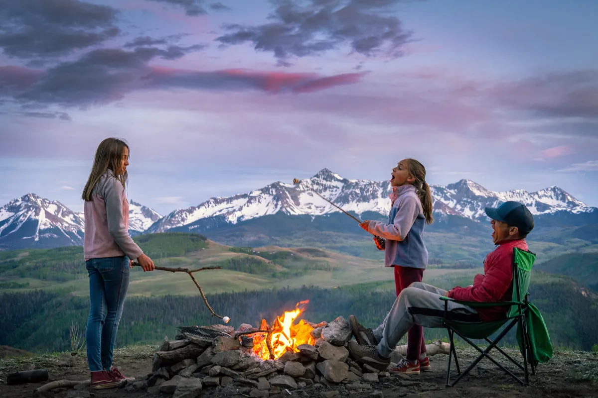 Children roast marshmallows over a campfire, surrounded by seating, with a dramatic mountain backdrop. The scene is lively and familial, emphasizing outdoor fun and togetherness.
